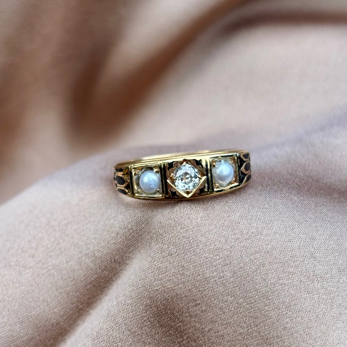 Gold ring with diamond centre and pearl stones black enamel on a beige fabric background